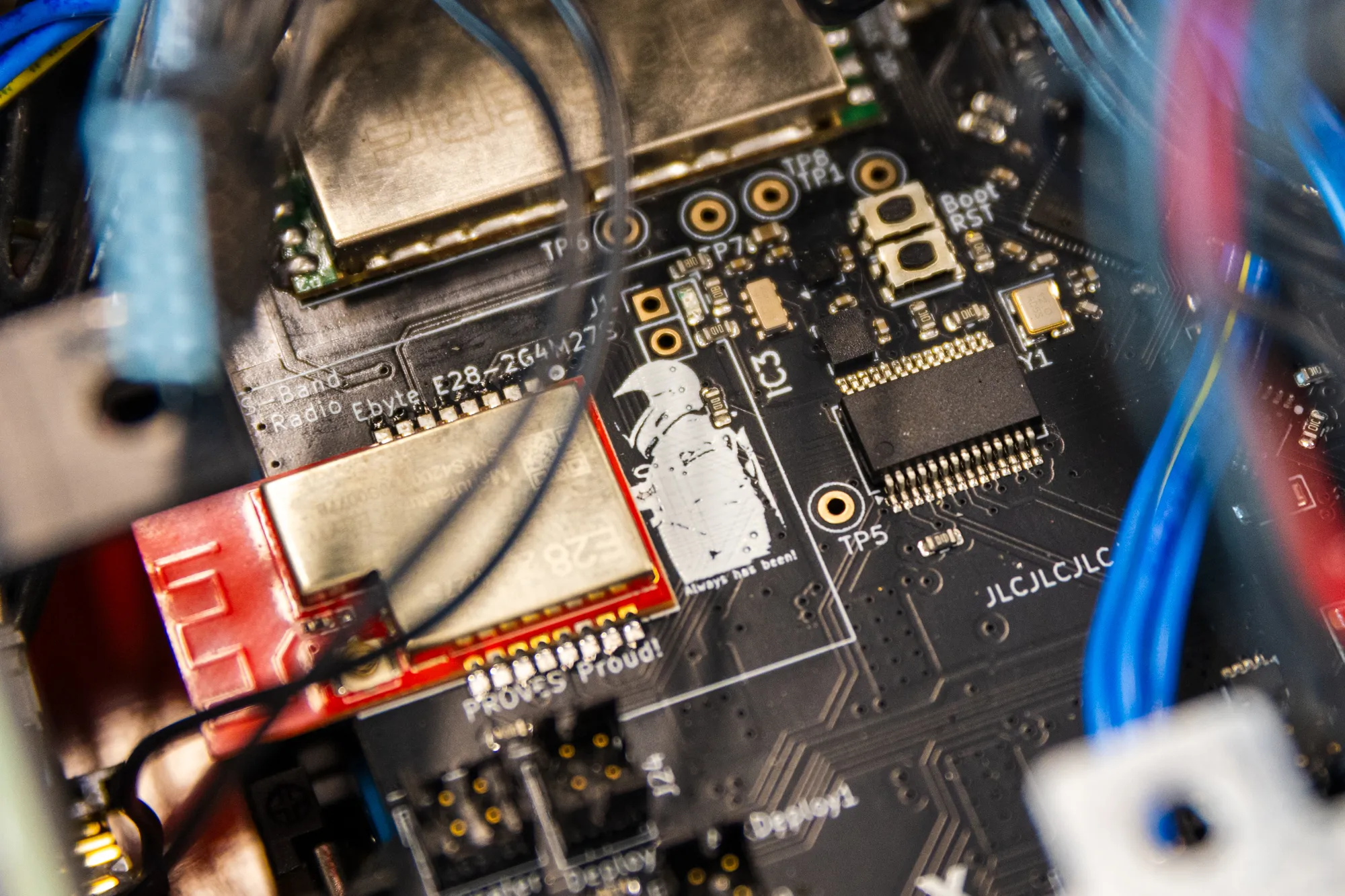 Close-up of a black circuit board with an S-band radio module and wiring in the foreground